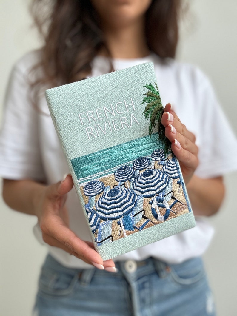 May include: A light blue clutch with a white embroidered design of a beach scene with palm trees, umbrellas, and lounge chairs. The text "French Riviera" is embroidered in white on the front of the clutch.