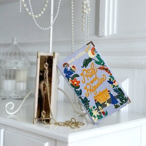 May include: A white clutch purse with a gold chain strap and a floral embroidered design. The clutch is open and the inside is lined with a light blue fabric. The clutch is sitting on a white surface. The text on the clutch reads "Jane Austen Pride and Prejudice".