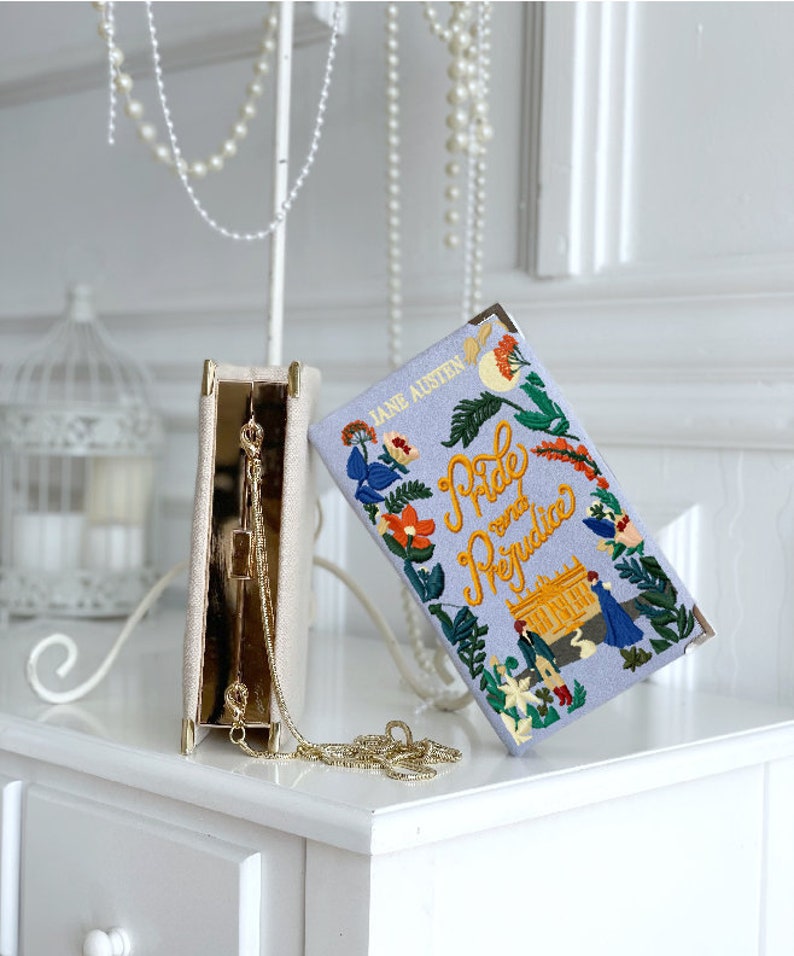 May include: A white clutch purse with a gold chain strap and a floral embroidered design. The clutch is open and the inside is lined with a light blue fabric. The clutch is sitting on a white surface. The text on the clutch reads "Jane Austen Pride and Prejudice".