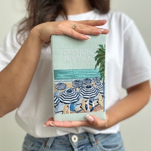 May include: A light blue clutch with a white embroidered design of a beach scene with palm trees and striped umbrellas. The text "FRENCH RIVIERA" is embroidered on the clutch.