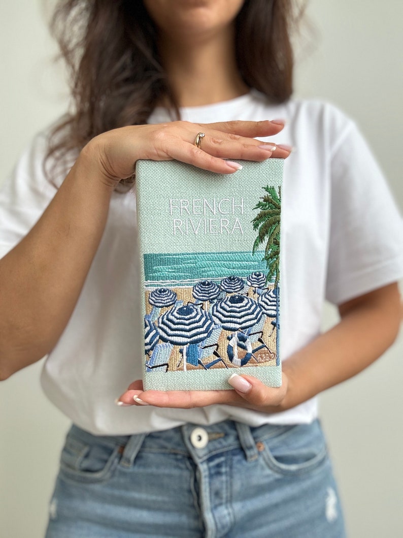 May include: A light blue clutch with a white embroidered design of a beach scene with palm trees and striped umbrellas. The text "FRENCH RIVIERA" is embroidered on the clutch.