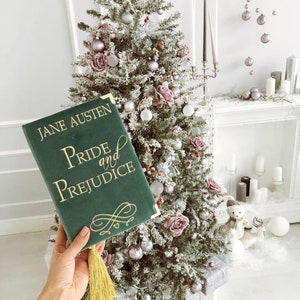 May include: A green velvet book with gold lettering that reads "Jane Austen Pride and Prejudice". The book has a gold tassel and is sitting in front of a Christmas tree decorated with pink and silver ornaments.