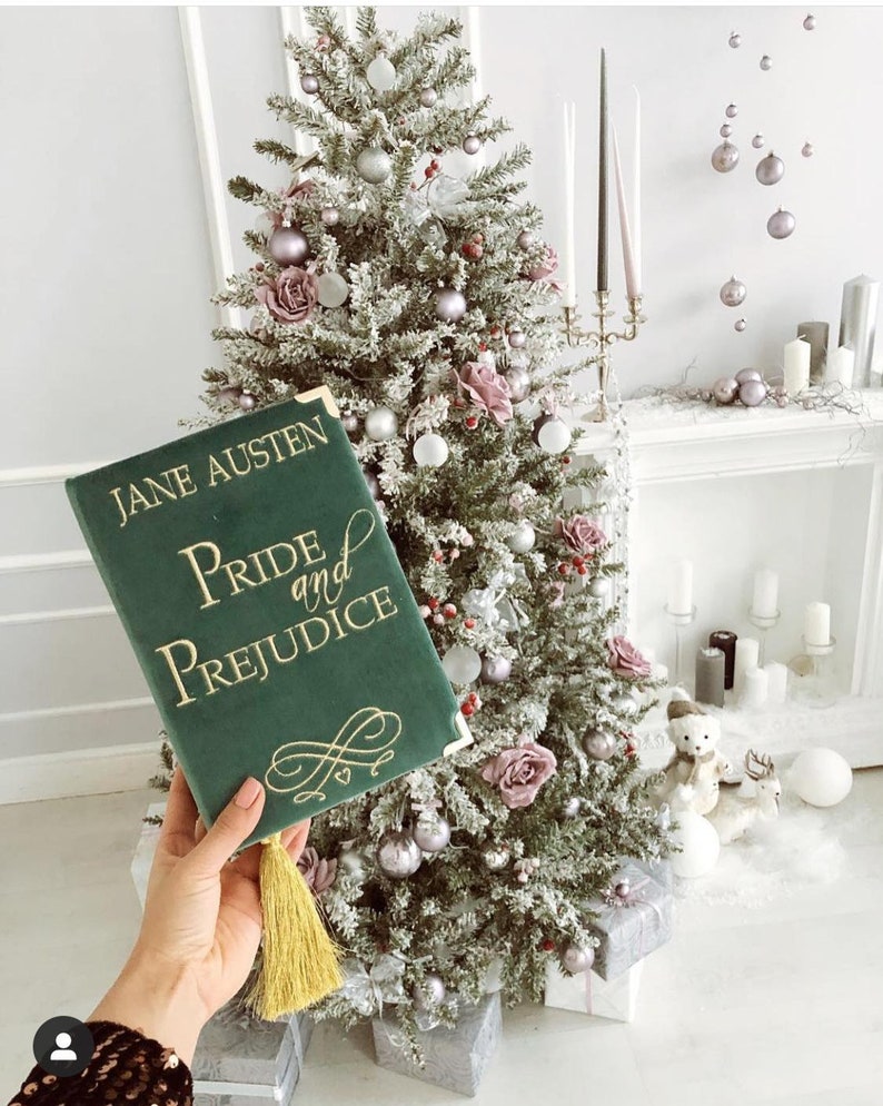 May include: A green velvet book with gold lettering that reads "Jane Austen Pride and Prejudice". The book has a gold tassel and is sitting in front of a Christmas tree decorated with pink and silver ornaments.