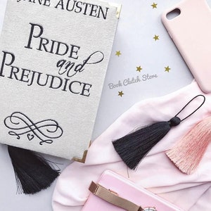 May include: A white book clutch with gold accents and black embroidery featuring the text "Jane Austen Pride and Prejudice". The clutch is decorated with a black tassel and is surrounded by pink fabric, a pink phone case, and gold stars.