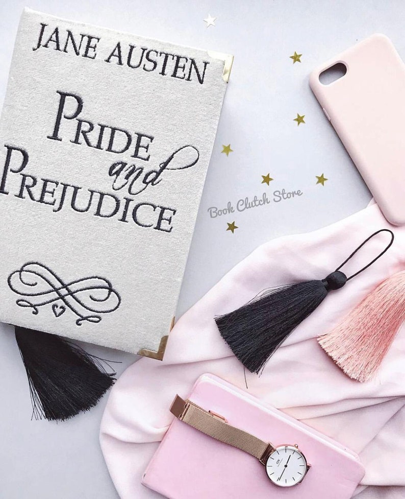 May include: A white book clutch with gold accents and black embroidery featuring the text "Jane Austen Pride and Prejudice". The clutch is decorated with a black tassel and is surrounded by pink fabric, a pink phone case, and gold stars.