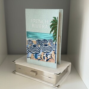 May include: A light blue clutch with a woven texture and an embroidered image of a beach scene with palm trees and striped umbrellas. The clutch has a silver metal clasp and the text "FRENCH RIVIERA" is embroidered on the front.