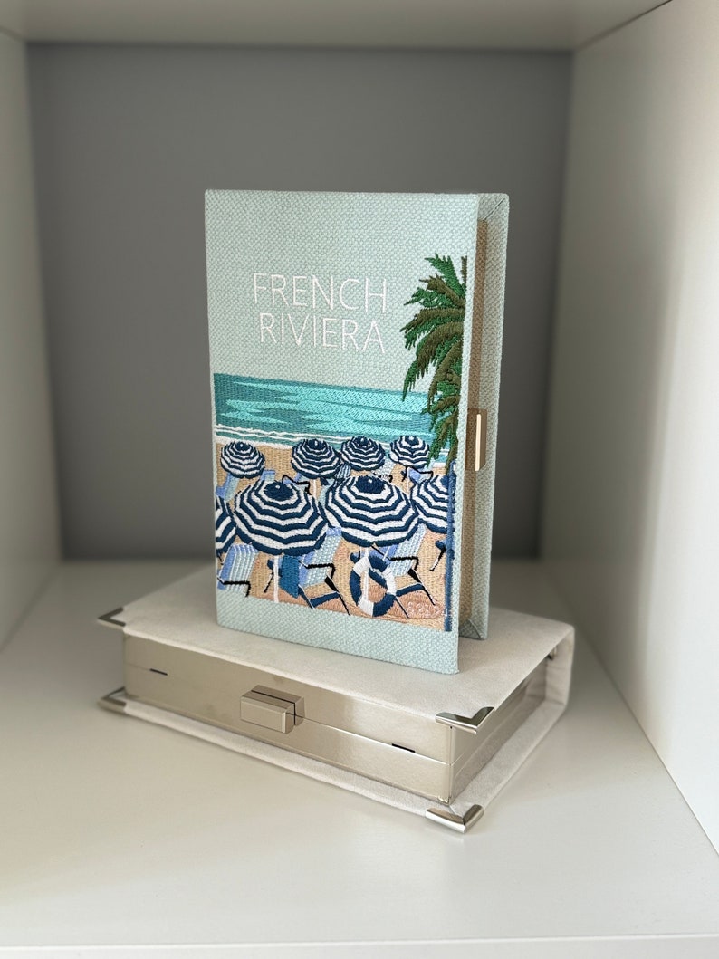 May include: A light blue clutch with a woven texture and an embroidered image of a beach scene with palm trees and striped umbrellas. The clutch has a silver metal clasp and the text "FRENCH RIVIERA" is embroidered on the front.