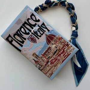 Pu&ograve; includere: Una pochette blu chiaro con un'immagine ricamata del Duomo di Firenze, Italia. La borsa ha una tracolla a fantasia blu e bianca con dettagli dorati.