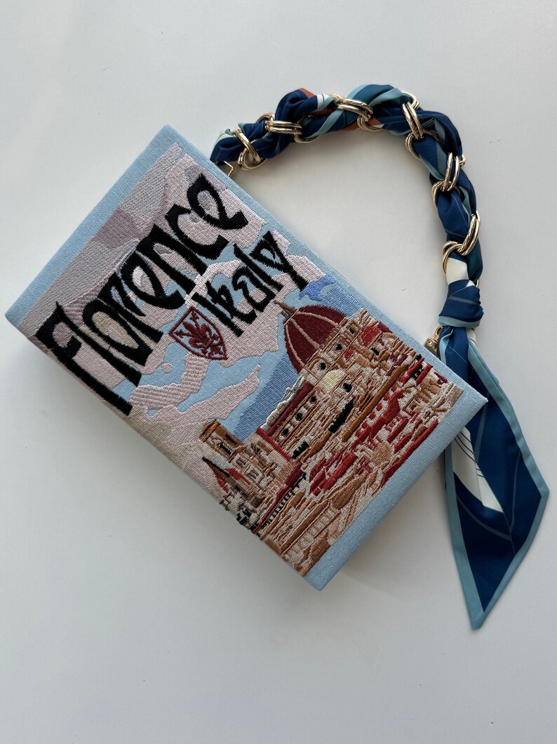 Pu&ograve; includere: Una pochette blu chiaro con un'immagine ricamata del Duomo di Firenze, Italia. La borsa ha una tracolla a fantasia blu e bianca con dettagli dorati.
