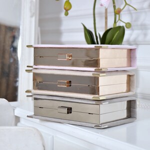 May include: Three stackable boxes with gold and silver metal frames and a soft, light pink, beige, and gray fabric lining. The boxes are stacked on a white dresser with a white drawer.