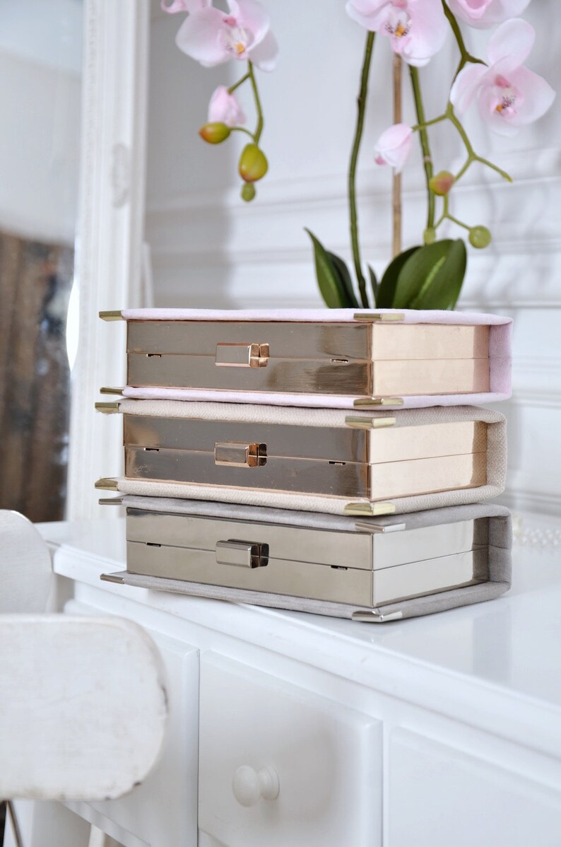 May include: Three stackable boxes with gold and silver metal frames and a soft, light pink, beige, and gray fabric lining. The boxes are stacked on a white dresser with a white drawer.