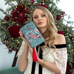 May include: A blue velvet clutch bag with a silver chain strap and a red tassel. The bag features an embroidered design of characters from the Nutcracker ballet, including a ballerina, a nutcracker soldier, and a mouse king. The text "The Nutcracker" is embroidered on the front of the bag.
