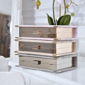 May include: Three stackable boxes with gold and silver metal frames and a soft, light pink, beige, and gray fabric lining. The boxes are stacked on a white dresser with a white drawer.
