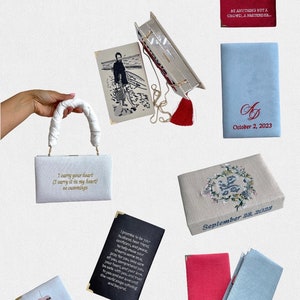 May include: A collection of small, white, fabric boxes with embroidered designs and text. The boxes have gold accents and some have tassels. The text on the boxes includes quotes, dates, and initials.