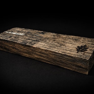 May include: A rectangular wooden block with a dark, weathered appearance. The wood grain is visible, showing shades of brown and gray. A small, dark leaf design is etched on one end. The block is set against a black background.