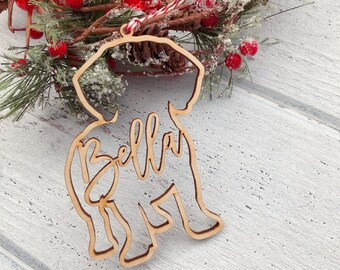 May include: A light-colored wooden ornament shaped like a dog silhouette with the name Bella cut out in cursive script. The ornament features a red and white string loop at the top for hanging and is displayed against a festive pine wreath.