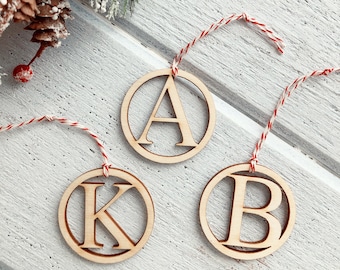 May include: Three wooden Christmas ornaments with the letters A, K, and B. Each letter is inside a circular frame and has a red and white string for hanging. The ornaments are on a white wooden surface, with a sprig of evergreen and berries in the background.