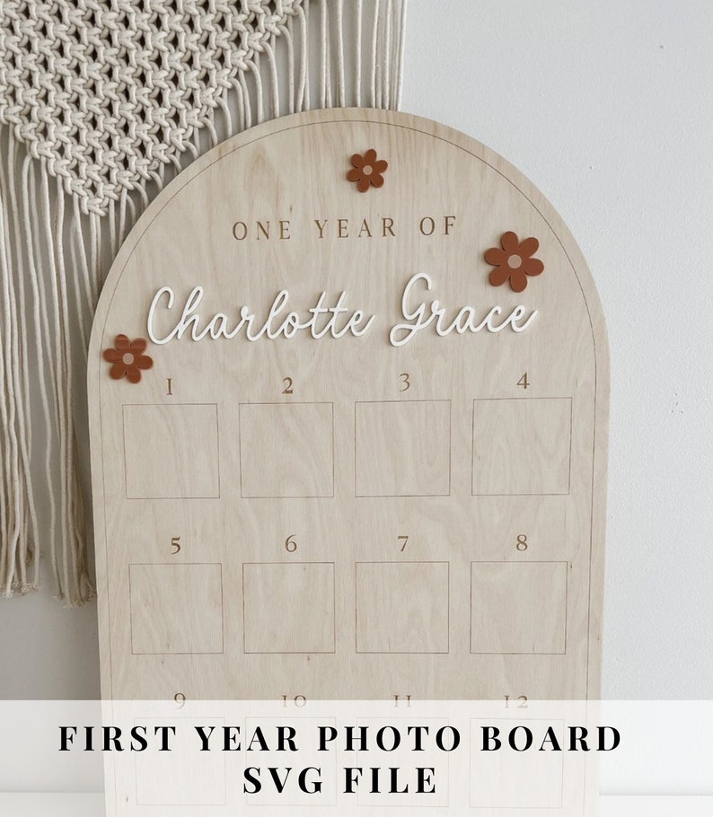 May include: A wooden photo board with a rounded arch top. The board has 12 squares for photos and the text "ONE YEAR OF Charlotte Grace" at the top. The board is decorated with small brown felt flowers.