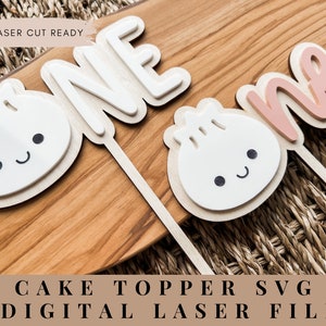 May include: A cake topper featuring the word "one" in white and the word "one" in pink, with two white cartoon dumplings with black eyes and smiles. The text "LASER CUT READY" is visible in the top left corner of the image. The text "CAKE TOPPER SVG DIGITAL LASER FILE" is visible at the bottom of the image.