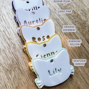May include: A collection of cat-shaped name tags in various colours, including purple, pink, orange, yellow, and seafoam green. Each tag features a name, with the colour names displayed. The tags are arranged on a wooden surface.