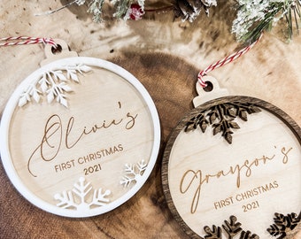 May include: Two wooden Christmas ornaments with snowflake cutouts. One has a white border and reads "Olivia's First Christmas 2021." The other has a dark brown border and reads "Grayson's First Christmas 2021." Both have red and white twine for hanging.