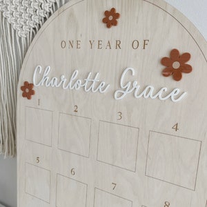 May include: A wooden arch-shaped growth chart with the text "ONE YEAR OF Charlotte Grace" and ten numbered squares for recording a child's height. The chart is decorated with brown felt flowers.