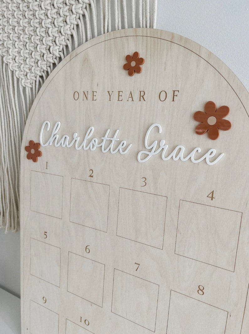 May include: A wooden arch-shaped growth chart with the text "ONE YEAR OF Charlotte Grace" and ten numbered squares for recording a child's height. The chart is decorated with brown felt flowers.