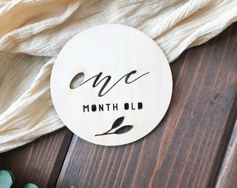 May include: A circular light-colored wooden plaque featuring the laser-cut text "one" in a cursive script, followed by the printed text "MONTH OLD" in a bold sans-serif font, with a small leaf graphic at the bottom.