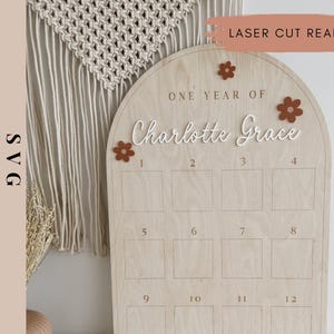 May include: A wooden arch-shaped sign with the text "One Year of Charlotte Grace" and twelve numbered squares for a child's first year growth chart. The sign is decorated with brown felt flowers.