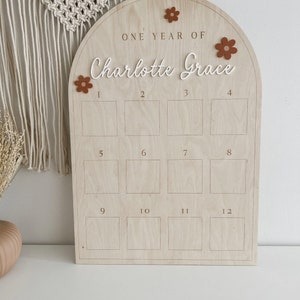 May include: A wooden arch-shaped board with the text "ONE YEAR OF Charlotte Grace" and 12 numbered squares. The board is decorated with brown felt flowers.