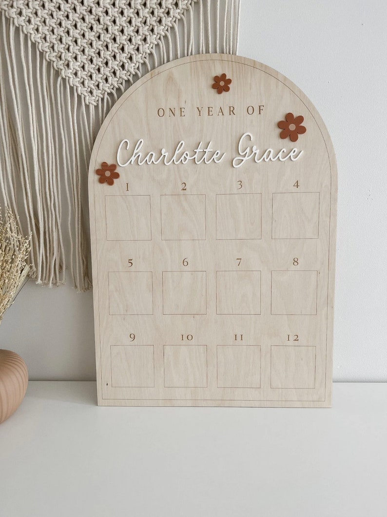 May include: A wooden arch-shaped board with the text "ONE YEAR OF Charlotte Grace" and 12 numbered squares. The board is decorated with brown felt flowers.