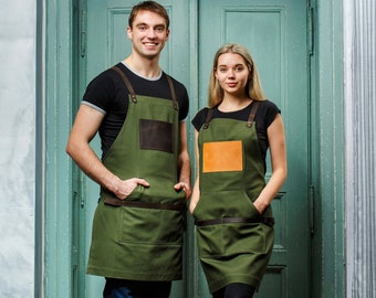 May include: Two people wearing green aprons with brown leather accents. The aprons have a large pocket on the front. The people are standing in front of a green door.