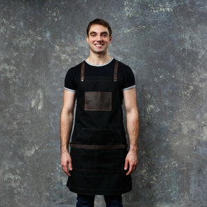 May include: A man wearing a black apron with brown leather straps and a brown leather patch pocket. He is smiling and looking at the camera.