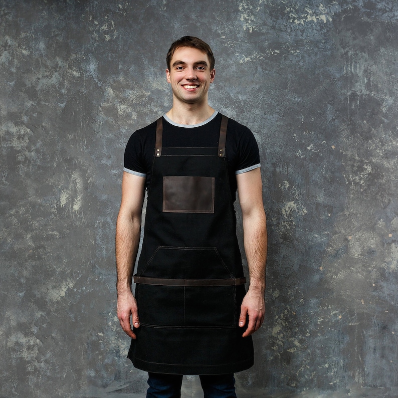 May include: A man wearing a black apron with brown leather straps and a brown leather patch pocket. He is smiling and looking at the camera.