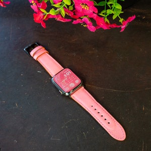 Puede incluir: Una correa de Apple Watch de cuero rosa con hebilla negra. La esfera del reloj muestra la hora 1:01.