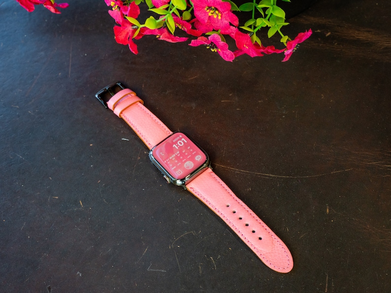 Puede incluir: Una correa de Apple Watch de cuero rosa con hebilla negra. La esfera del reloj muestra la hora 1:01.