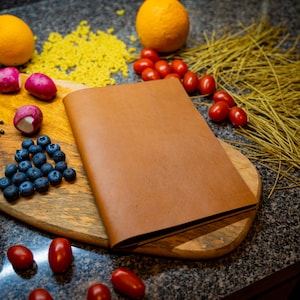 May include: A brown leather-bound journal or notebook on a wooden cutting board surrounded by various food items, including blueberries, radishes, tomatoes, pasta, and oranges.