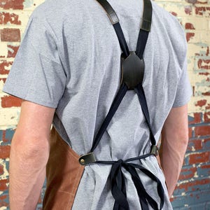 May include: A brown leather apron with adjustable black straps and a black leather buckle. The straps cross in the back and tie at the waist.