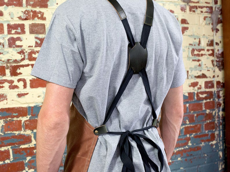 May include: A brown leather apron with adjustable black straps and a black leather buckle. The straps cross in the back and tie at the waist.