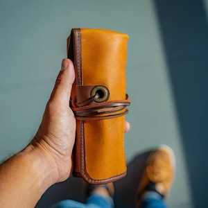 May include: A brown leather pencil case with a brown leather strap and a brass buckle. The case is rolled up and held in a hand.