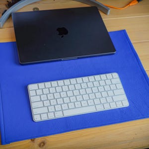Könnte beinhalten: Eine rechteckige blaue Schreibtischunterlage mit strukturierter Oberfläche, auf der ein geschlossener dunkelgrauer Laptop mit Apple-Logo und eine weiße kabellose Tastatur liegen. Die Unterlage hat genähte Kanten und eine einheitliche blaue Farbe.