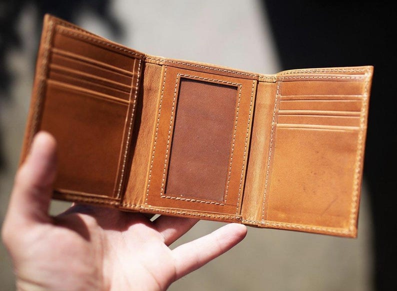 May include: Open, light brown leather trifold wallet. The wallet has multiple card slots and a clear ID window. The interior is a darker shade of brown. The edges are stitched with a lighter thread. The wallet is open, displaying its interior.