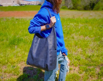 May include: A person wearing a blue hoodie and jeans is carrying a large, dark blue leather tote bag with a long strap.