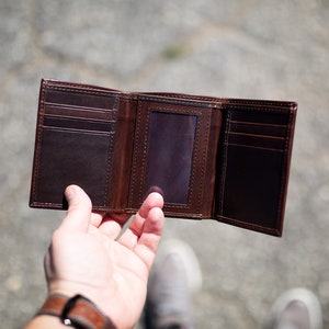 May include: Open, brown leather trifold wallet with multiple card slots and an ID window. The wallet is displayed open, showcasing its interior design and construction. The leather has a rich, dark brown color.
