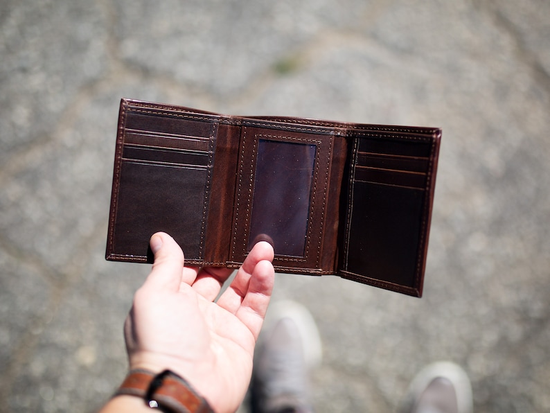 May include: Open, brown leather trifold wallet with multiple card slots and an ID window. The wallet is displayed open, showcasing its interior design and construction. The leather has a rich, dark brown color.