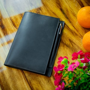 May include: A black leather journal with a silver pen tucked inside. The journal is lying on a wooden table with two oranges and a bouquet of pink flowers.