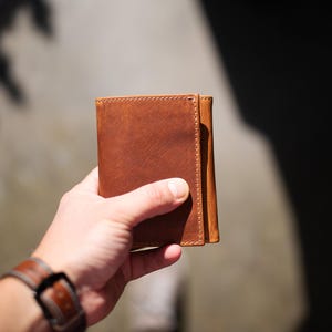May include: A brown leather wallet with visible stitching is held in a hand. The wallet is a tri-fold design, and the leather has a natural, textured appearance. A brown leather watch with a buckle is visible in the foreground.