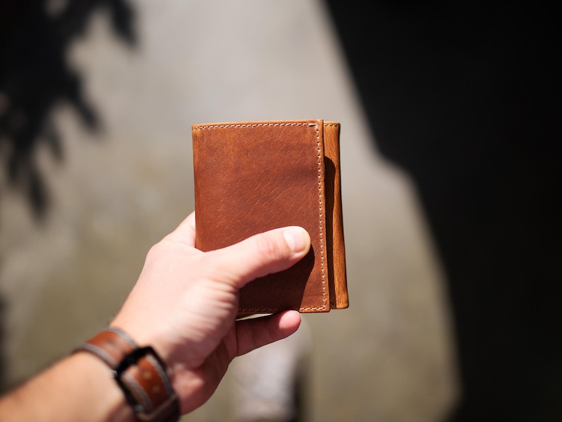 May include: A brown leather wallet with visible stitching is held in a hand. The wallet is a tri-fold design, and the leather has a natural, textured appearance. A brown leather watch with a buckle is visible in the foreground.