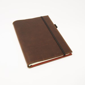 Puede incluir: Un cuaderno de cuero marrón con cierre elástico negro y lomo rojo.