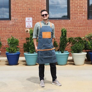 May include: A man wearing a blue denim apron with brown leather straps and a brown leather patch pocket. He is standing in front of a brick wall with potted plants.